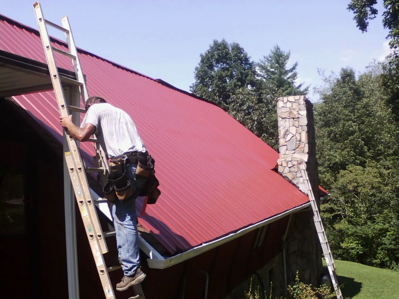 Roofer on a ladder beside a red metal roof and stone chimney — Soffit & Fascia Repair by Roofing Brothers in Van